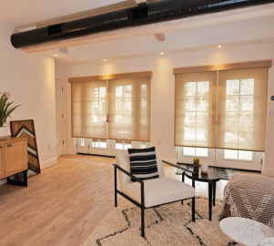 A modern living room with wooden floors, a white armchair, a rug, and large beige blinds adding a touch of colour to the windows. A black ceiling beam is visible, and a cabinet with decorative items sits elegantly in the corner.