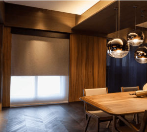 Dining room with wood-paneled walls, a wooden table, beige chairs, metallic pendant lights, and a large window partially covered by fabric blinds that add a subtle touch of color.