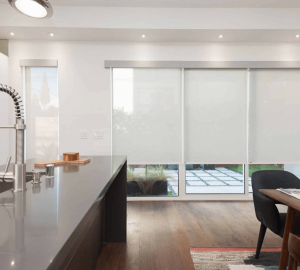 Modern kitchen with a sleek countertop and sink on the left. Large windows with white blinds add a touch of color to the back wall. A dark chair and part of a table are on the right.