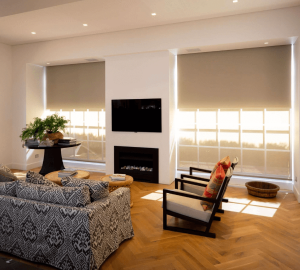 A living room with a black TV, fireplace, patterned sofa, two armchairs, and a small table with a plant basks in filtered sunlight. The large windows feature closed blinds that add to the ambiance and subtly enhance the colour of the decor.