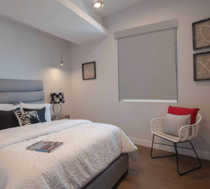 A modern bedroom featuring a neatly made bed with white and gray bedding, complemented by a side table with a lamp. A white chair adorned with red and striped cushions adds a splash of colour, while the room's aesthetics are softened by the closed window blinds showcasing elegant wall art.