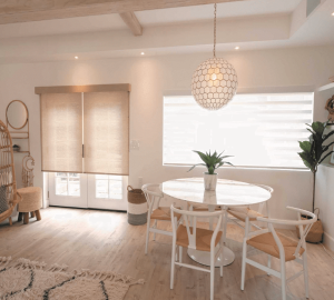 A bright dining area features a round table with four chairs and a potted plant centerpiece. A spherical pendant light complements the space, while large windows with fabric blinds control the influx of natural light.