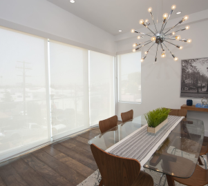 A dining room featuring a sleek glass table and wooden chairs, illuminated by a modern chandelier. Large windows with stylish blinds add to the ambiance, while a splash of color comes from a framed picture on the wall.