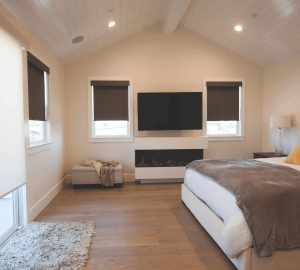 A modern bedroom features a large bed, TV on the wall, and a cozy fireplace. Two windows with dark fabric blinds complement the light-colored walls, while wooden floors and a rug add warmth. A small bench by one window completes this harmonious blend of texture and color.