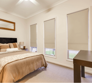 A bedroom features a bed, nightstand, and three tall windows fitted with fabric blinds. Beige tones dominate the décor, while a wooden-framed mirror elegantly hangs above the headboard, enhancing the room's serene ambiance.