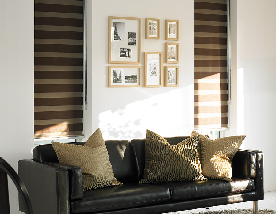Against a white wall with framed pictures, a black leather sofa with patterned cushions stands. Two windows adorned with brown Zebra Blind Range B flank the scene, adding color and elegance to the ambiance.