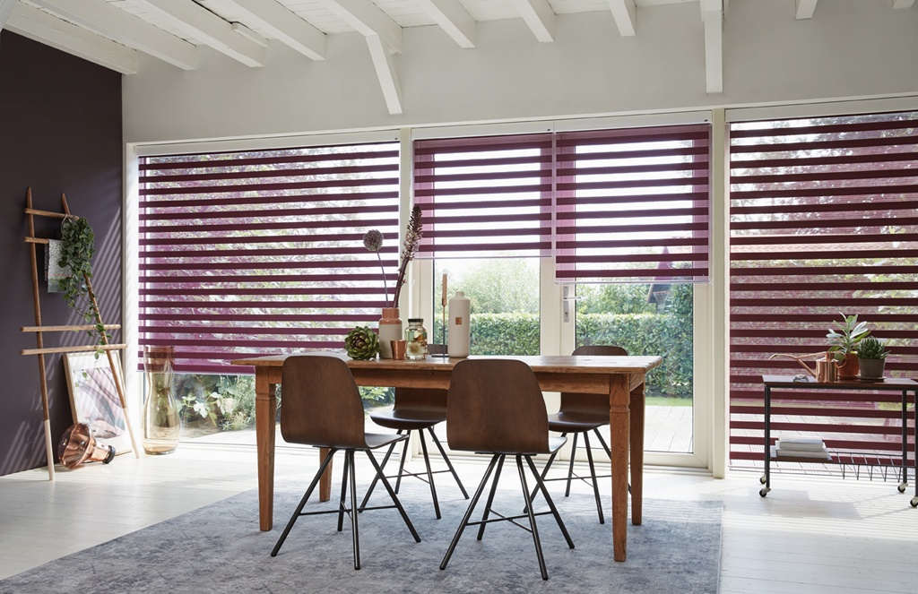 A dining room featuring a wooden table with four chairs is complemented by Zebra Blind Range B on large windows. Lush plants and tasteful decor adorn the space, adding harmony and invitation.