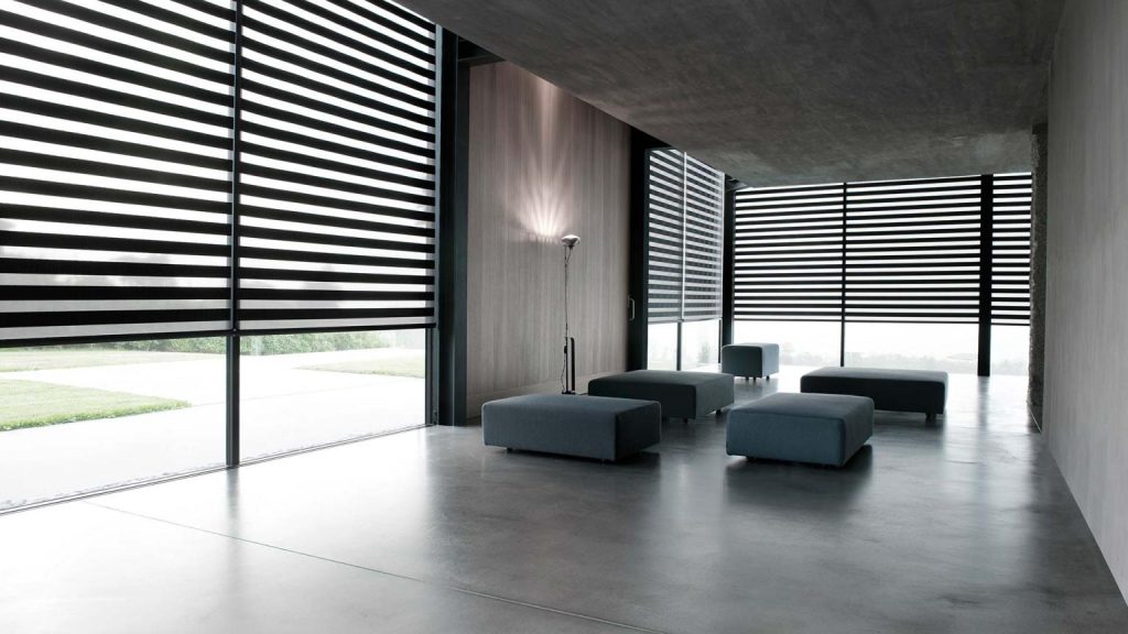 A modern interior with large windows features the Zebra Blind Range B, minimalist gray ottomans, and a floor lamp on a sleek concrete floor. The subtle play of light and shadow enhances the elegant ambiance with gentle touches of color.