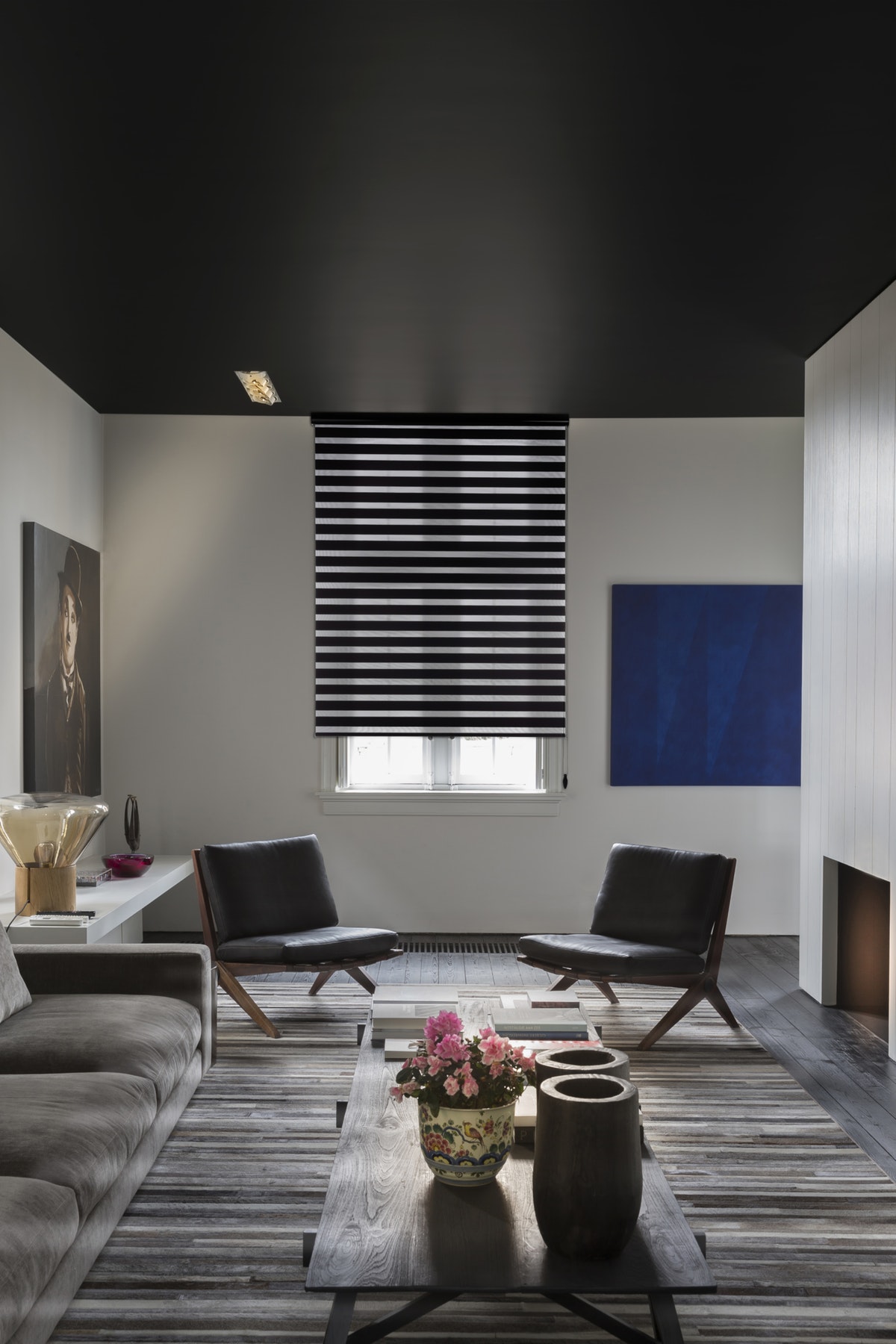Modern living room featuring a gray sofa, two black chairs, and Zebra Blind Range B. The crisp white walls display a portrait and an abstract painting. Decorative items are elegantly arranged on the table and floor.