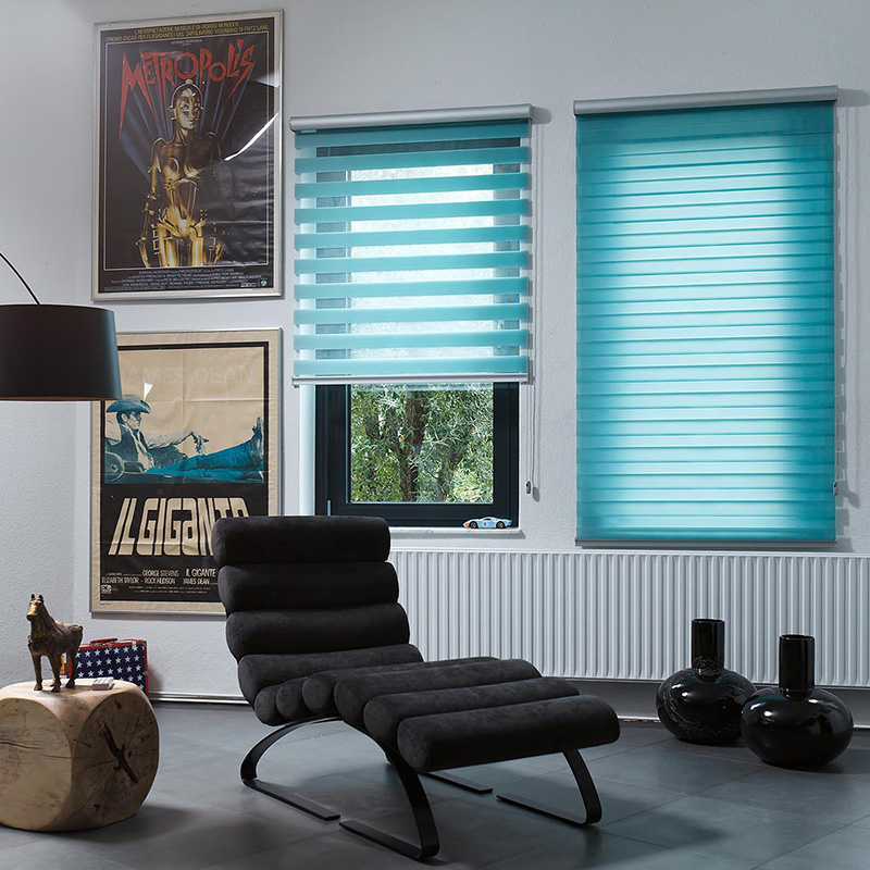 A room featuring two Zebra Blind Range B blue-striped blinds adds a splash of color, complemented by a black lounge chair, decorative posters, and a floor lamp. Two black vases rest on the radiator, while a wooden stool supports an elegant horse figurine.