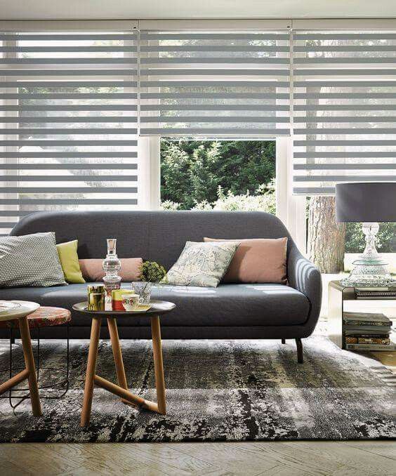A modern living room features a gray sofa, assorted cushions, a patterned rug, and a round coffee table. Large windows with Zebra Blind Range B in bold stripes reveal lush greenery outside.