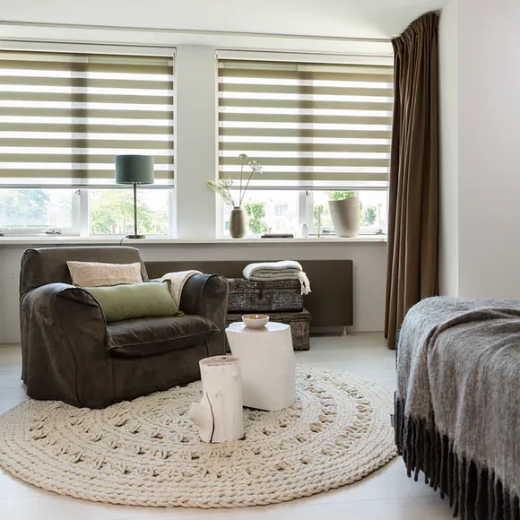 Cozy room with a large armchair, round woven rug, side table, stacked blankets by the window, and soft curtains gently filtering light through Zebra Blind Range B. In the foreground is a bed draped with a gray blanket.