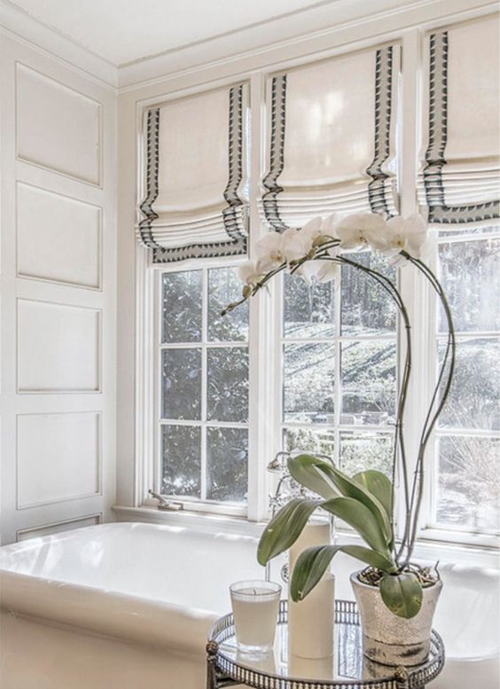 A bright bathroom features a white bathtub and large windows adorned with elegant Roman shades, adding a touch of color. An orchid sits on a small round table next to a glass of water, completing the serene setting.