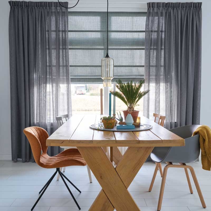 A cozy dining room featuring a wooden table set for four, adorned with a centerpiece of plants and candles. Large windows dressed with gray curtains add warmth, while keeping the space open to light.