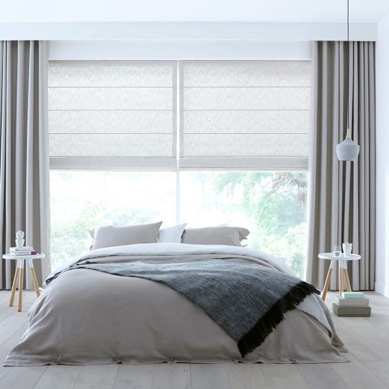 A minimalist bedroom features a neatly made bed with a gray blanket, two side tables, and a large window adorned with Roman shades and fabric curtains that add a touch of color.