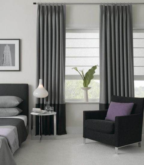 A modern bedroom features gray fabric curtains, a sleek black armchair adorned with a purple cushion, a side table topped with a stylish lamp, and a cozy bed draped in gray bedding. A framed picture decorates the wall, adding to the room's elegant charm.