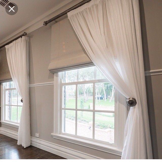 Large window with white sheer curtains tied back, revealing beige blinds. The window offers a view of greenery outside. The room's light gray walls complement the dark wood flooring, creating a harmonious blend of colors.