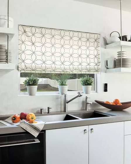 Modern kitchen with a sink, dual faucets, and colorful fabric blinds on the window. Potted plants line the sill, and dishes are neatly arranged on shelves. A bowl of fruit sits invitingly on the counter.