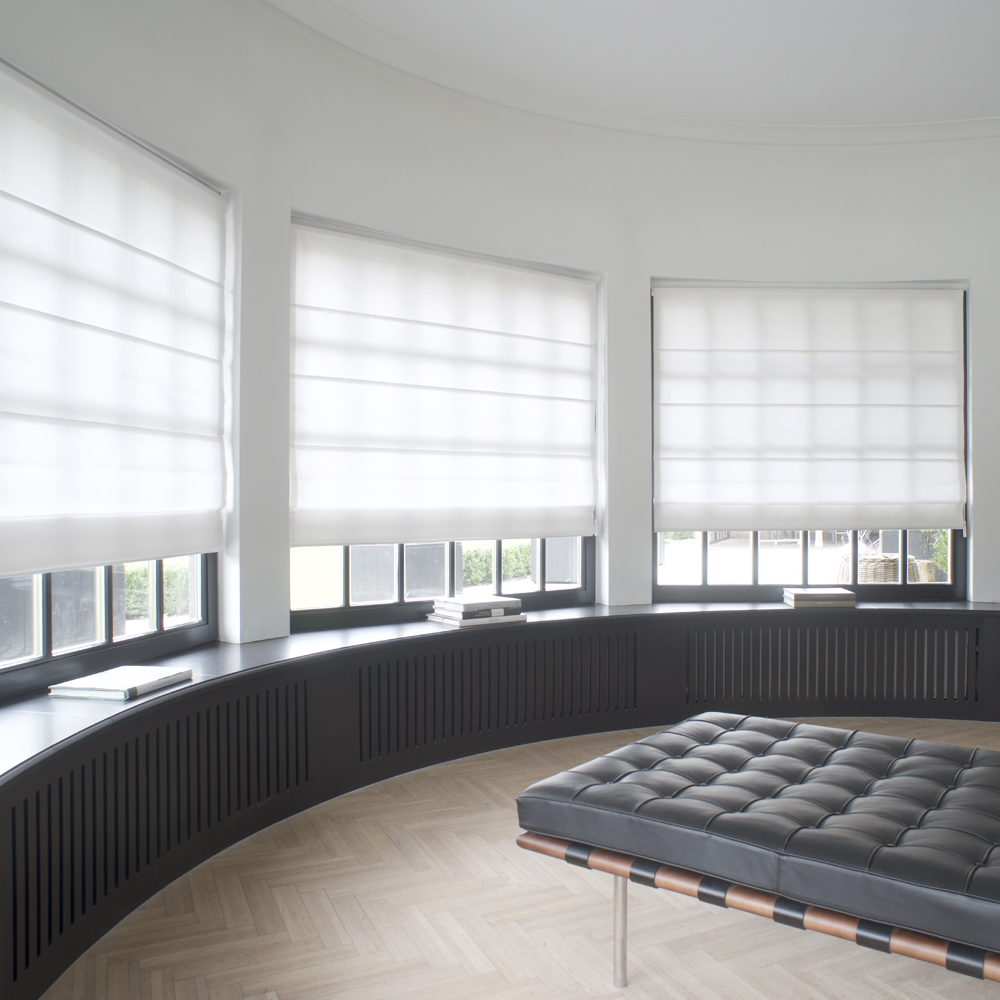A modern room with large curved windows featuring white Roman fabric shades, a black bench, and a light wood herringbone floor creates an inviting and stylish atmosphere.