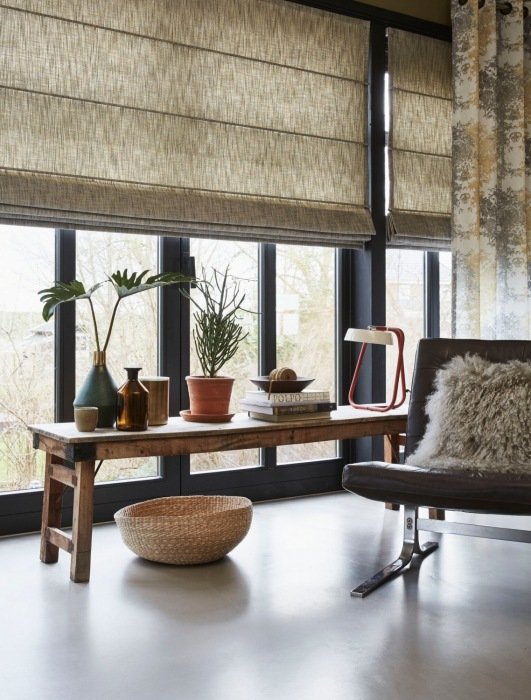 A room with large windows adorned with colorful Roman shades, a wooden table hosting potted plants, a lamp, decorative items, and a chair with a fur cushion next to a wicker basket on the floor.