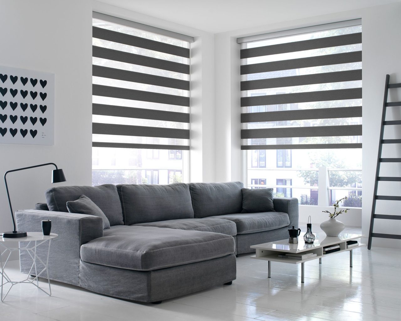 A modern living room features a gray sectional sofa and warm-colored Zebra Blind Range B. Minimalist decor includes a ladder shelf in the corner, paired with small tables and decorative items.