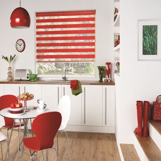 A modern kitchen showcases white cabinets with vibrant red accents and a round table surrounded by red and white chairs. The window features Zebra Blind Range B, while decor items such as red boots and a plant picture provide splashes of color.