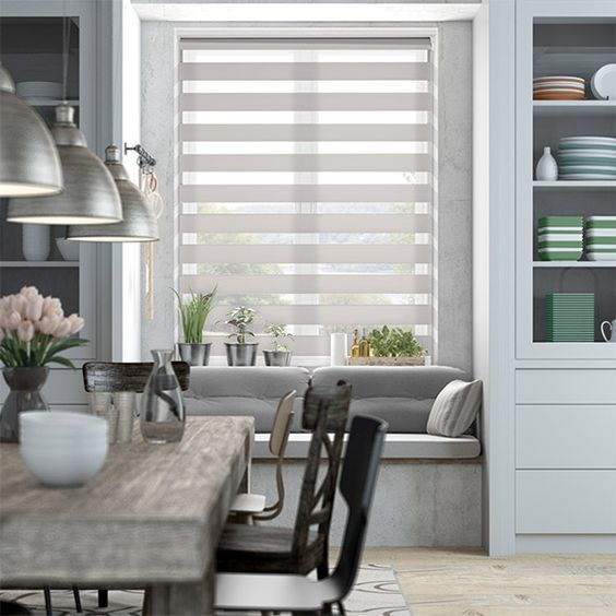 Modern dining area featuring a wooden table, metal chairs, and pendant lights. A cozy window seat with cushions and plants sits beside a window adorned with Zebra Blind Range B. Shelves display a vibrant array of colorful dishes.