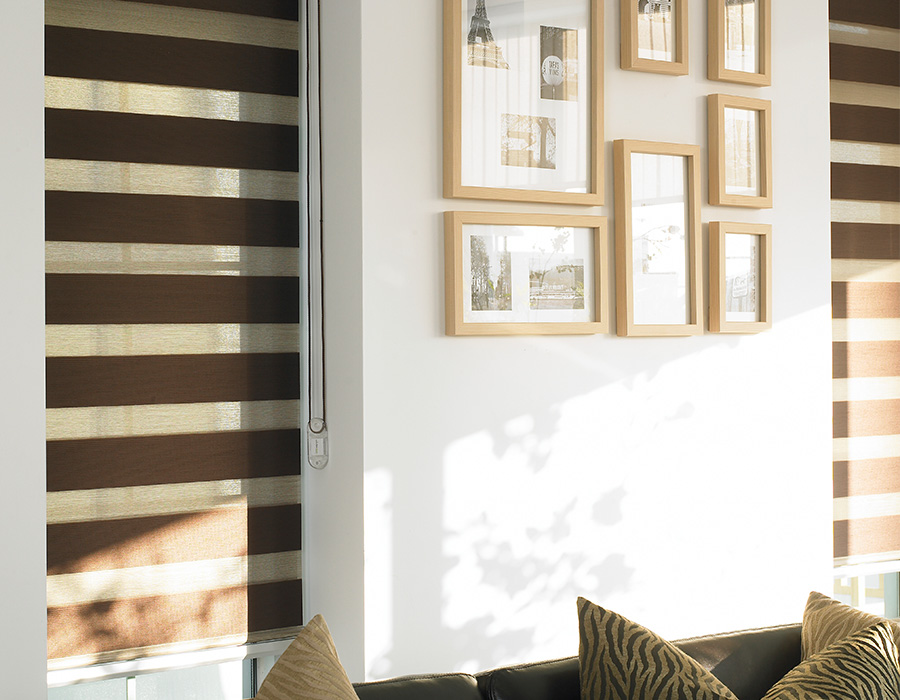 A room featuring Zebra Blind Range B casts a warm glow over the black sofa accented with patterned cushions, while framed pictures elegantly adorn the wall.