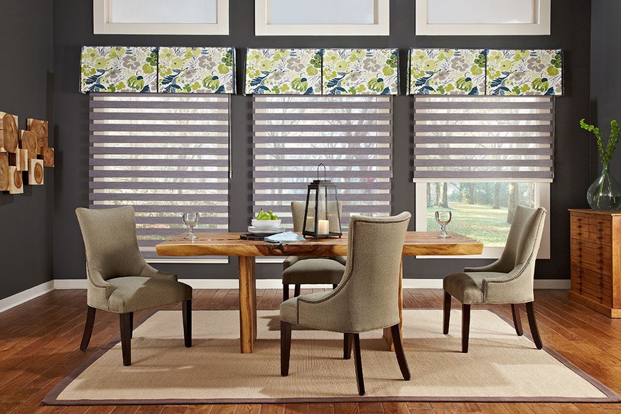 Dining room featuring four upholstered chairs around a wooden table, framed by large windows adorned with Zebra Blind Range B blinds and floral valances. Earth-toned decor and wooden accents complete the setting.