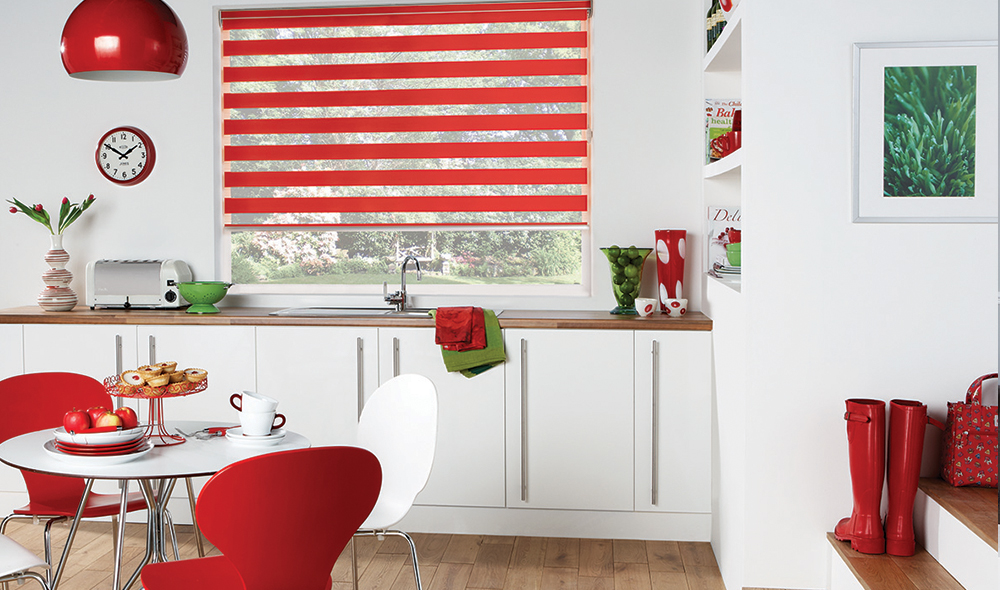 A modern kitchen with red and white decor, featuring a round table, chairs, vibrant Zebra Blind Range B in red, a wall clock, boots, and various kitchen items.