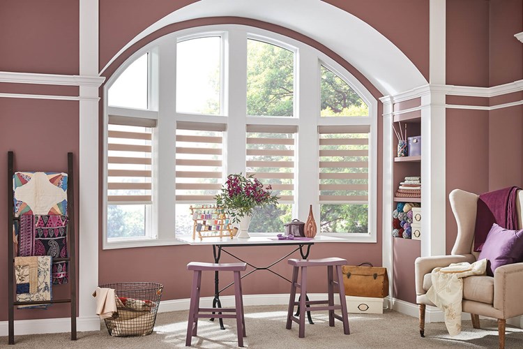 A room with mauve walls features an arched window adorned with Zebra Blind Range B, two matching mauve stools, a small table displaying books and flowers, and a ladder draped with cozy quilts.