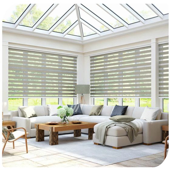 A spacious, sunlit living room features a large sectional sofa and wooden coffee table beneath a stunning glass ceiling. Surrounded by tall windows with Zebra Blind Range B blinds, this inviting space basks in natural light.