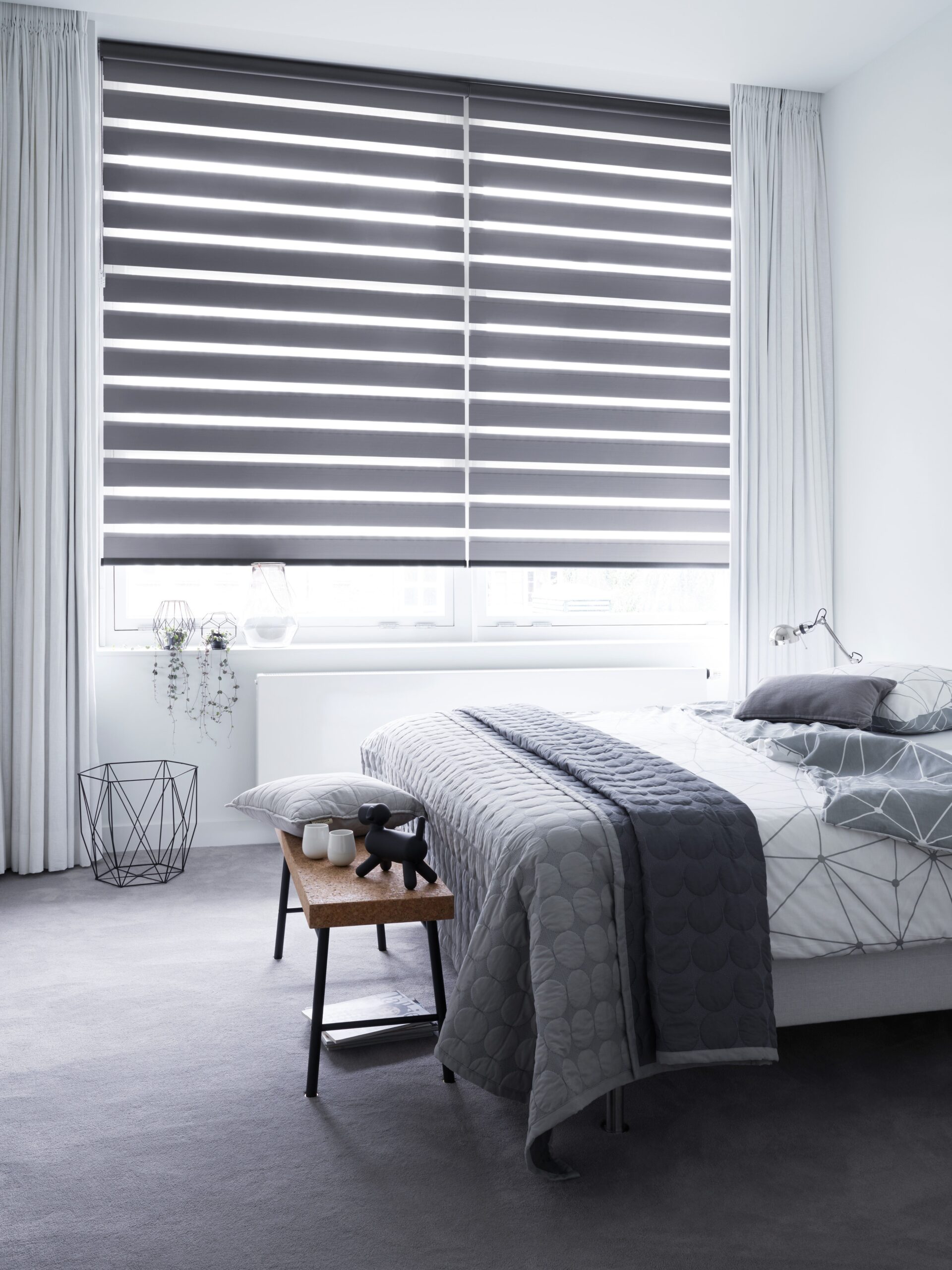 A modern bedroom showcases a neatly made bed with gray bedding and Zebra Blind Range B. White fabric curtains enhance the small bedside table adorned with decor items, while light gray walls and floors complete the serene ambiance.