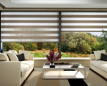 Modern living room with two white sofas and a glass coffee table adorned with flowers. Large windows, dressed in Zebra Blind Range B, provide a picturesque view of the wooded landscape, adding color to the serene setting.