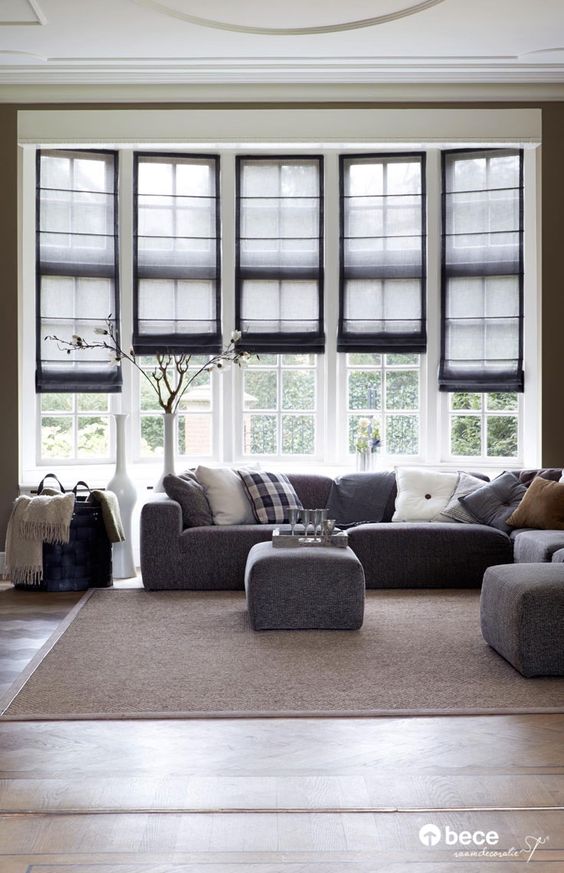 A living room with a grey sofa, ottoman, and a tall white vase. Large windows feature black-framed Roman blinds behind. A light brown rug adds a touch of warmth to the wooden floor.
