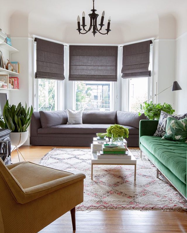 Bright living room with a gray sectional, green couch, and beige armchair. Rugs cover wooden floors while Roman Blind Range A filters the light. Plants decorate the space, and a black chandelier hangs from the ceiling.