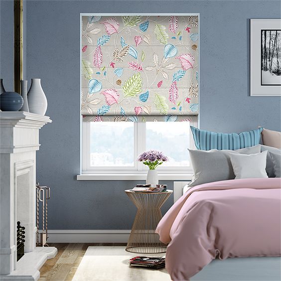 A bedroom showcasing a bed and side table is enhanced by Roman Blind Range A, featuring a colorful leaf pattern, adding charm beneath the white-framed pictures on the serene blue wall.