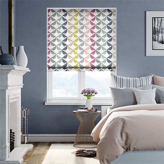 A bedroom showcasing a bed and a round side table with flowers, accented by colorful blinds on the window. The blue walls hold framed art above a crisp white fireplace, adding to the room's charming allure.
