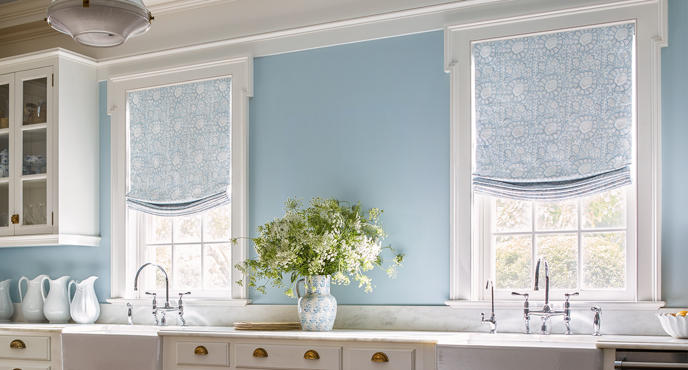 A kitchen with pale blue walls features two windows adorned with Roman Blind Range A patterned in blue, a white countertop with floral arrangements, double sinks, and white cabinetry.