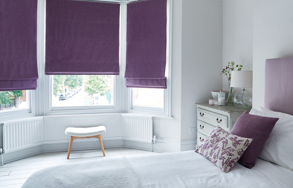 A bedroom showcasing a white bed with purple pillows and vases on the dresser, features three windows adorned with Roman Blind Range A in purple fabric and a cozy bench nearby.