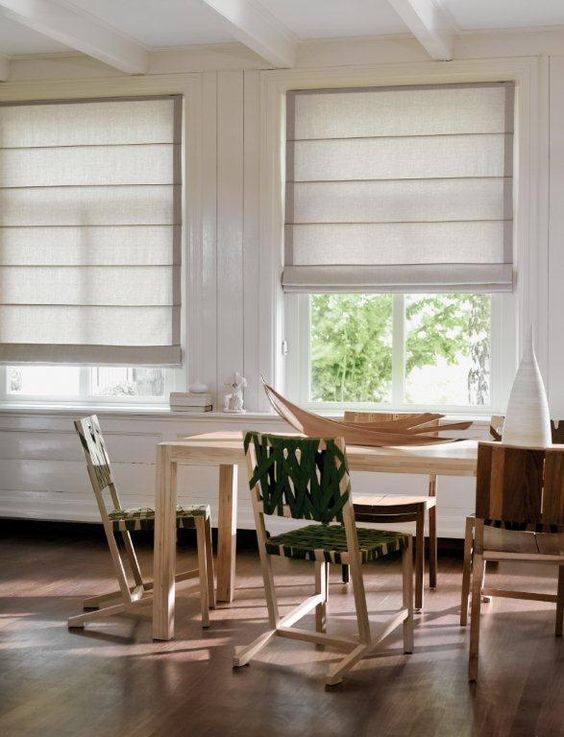 The dining area features a wooden table and chairs, large windows adorned with beige Roman Blind Range A fabric shades, and white walls. Natural light streams in, illuminating the room.