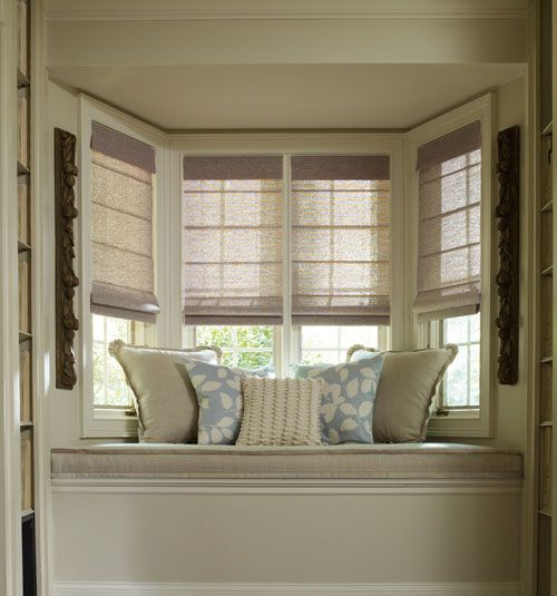 A cozy window seat with beige cushions and patterned pillows is framed by Roman Blind Range A on three bay windows in a softly lit room, where the blend of fabric hues adds warmth.