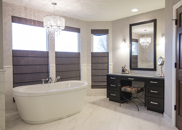 Spacious bathroom with a freestanding bathtub, sleek black vanity with mirror, and decorative chair. Large windows feature Roman Blind Range A in gray fabric, while chandelier lights warm the room in a subtle glow.