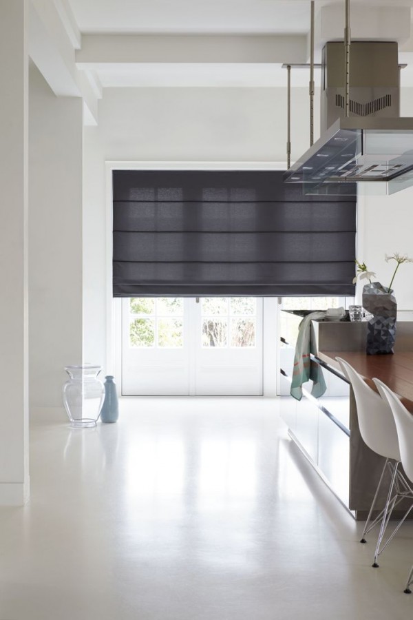 A modern kitchen features a glossy island, white chairs, and sleek Roman Blinds from Range A covering large windows. Minimalist decor includes a clear vase on the floor and a jacket draped over a chair.