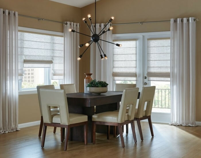 The dining room features a dark wooden table and six beige chairs, enhanced by sheer curtains and Roman shades that subtly filter light. A central black chandelier adds elegance, while the brown walls and wood floor provide warmth, seamlessly blending color with fabric in every detail.