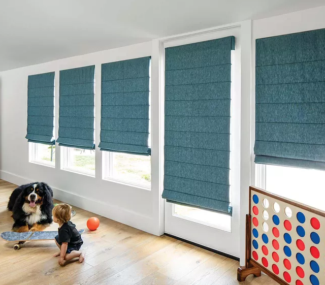 A child plays with a skateboard next to a large dog in a room adorned with Roman Blind Range A in blue fabric and a Connect Four game.
