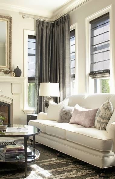 A living room with a white sofa, patterned cushions, and a round coffee table. Fabric drapes complement the dark curtains by the tall windows, while a colourful lamp adds vibrancy. A fireplace is visible on the left.