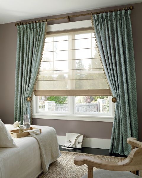 Bedroom window with teal curtains and beige Roman shades. A bed with white linens and a side table with decorative items are visible. Outside, a glimpse of greenery and a fence can be seen through the blinds, adding a touch of color to the serene setting.