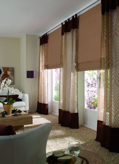 The living room features large windows adorned with brown and sheer patterned fabric curtains, emitting a soft light that enhances the colour of the beige carpet. White furniture complements a wooden coffee table with plants, while a purple floor lamp adds a vibrant touch.