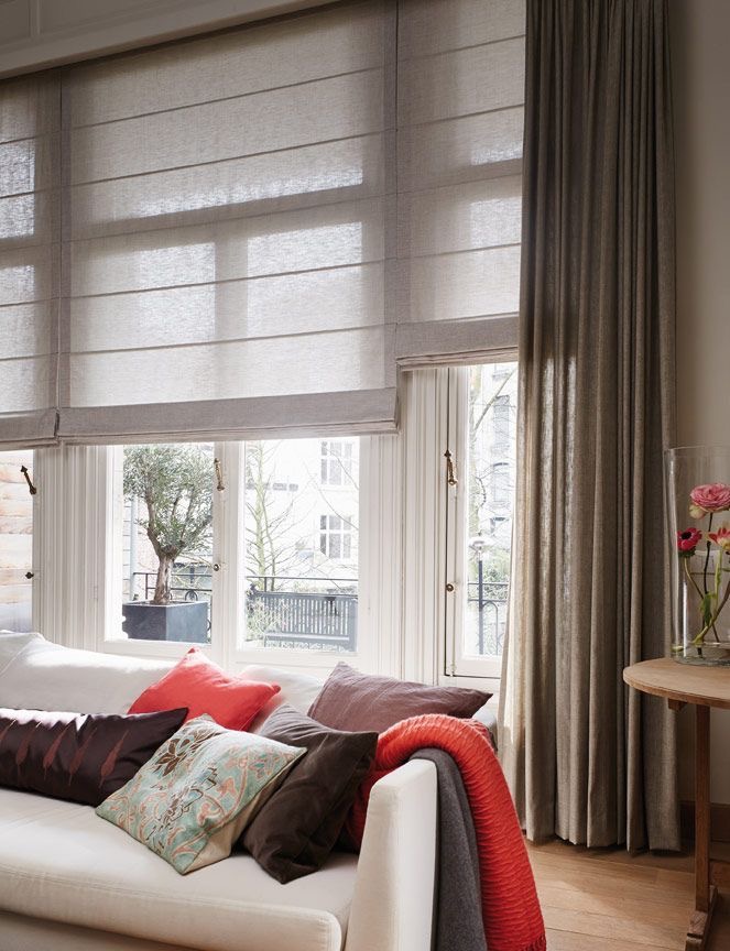 A living room with a white sofa, colorful cushions, and a red throw blanket. Large windows feature sheer Roman shades and long fabric curtains. A round wooden table holds a glass vase with flowers.